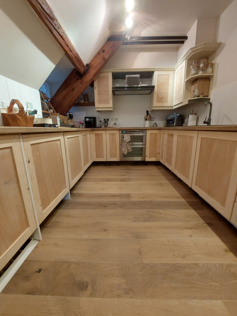 A spacious kitchen featuring wooden cabinets, a large window, and wooden flooring, with a sloped ceiling and exposed beams.