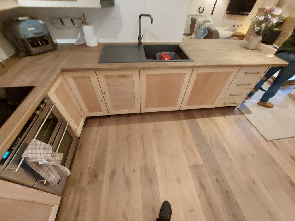 A modern kitchen featuring wooden cabinetry, a dark sink, and a stove with an oven. There is also a dish towel hanging from the oven handle and a person standing nearby.