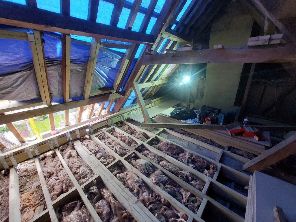 Interior view of an attic under construction, showing exposed wooden beams, insulation, and a blue tarp covering the roof structure. A light source illuminates the space.
