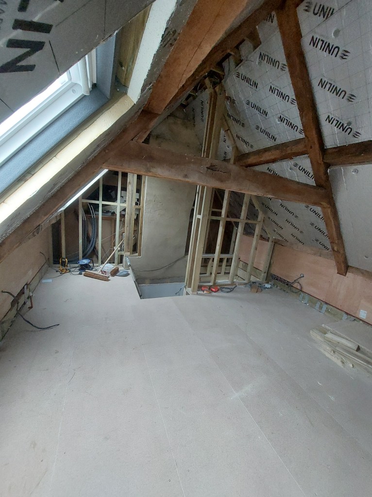 Installation view of an unfinished attic space, showcasing exposed beams, insulation material, and construction tools scattered on the floor.