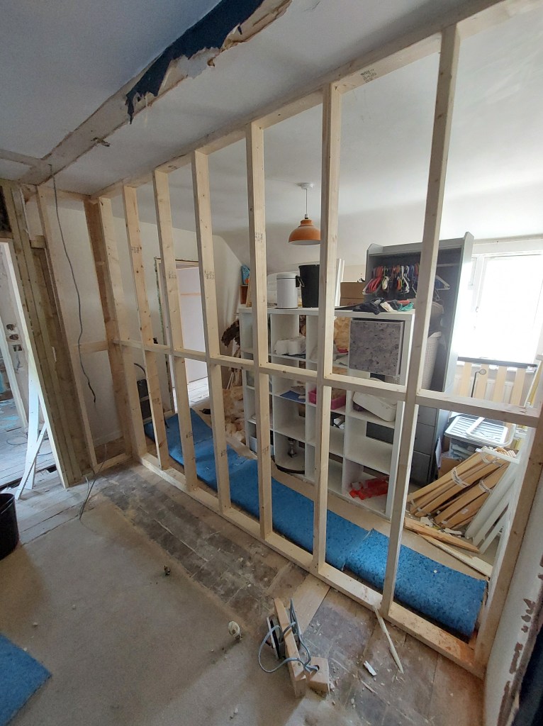A partially constructed interior wall with wooden framing, showing part of a room with shelves and a blue carpet underneath.