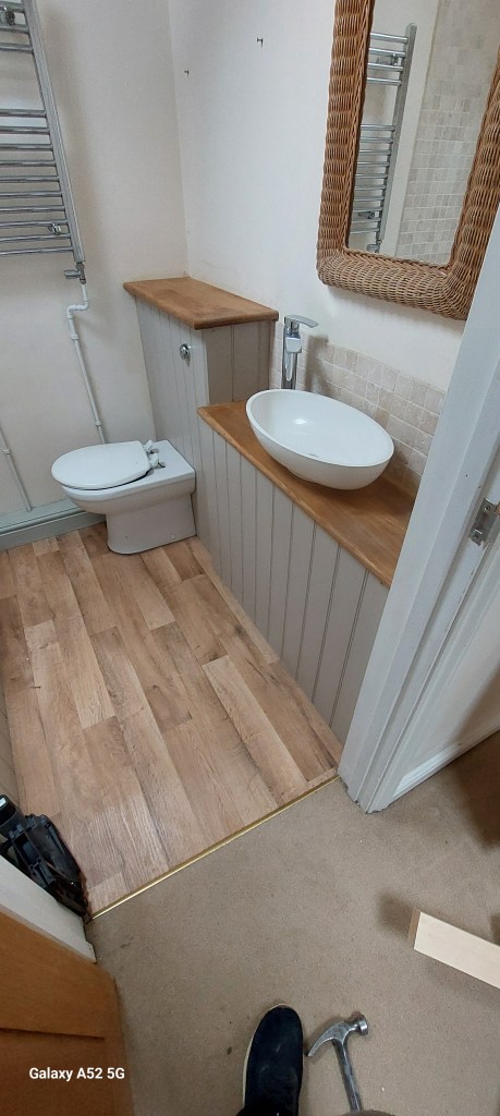 A modern bathroom featuring a white basin, a toilet, wooden countertops, and a large mirror. The flooring is a light wood, and there is a towel rack on the wall.