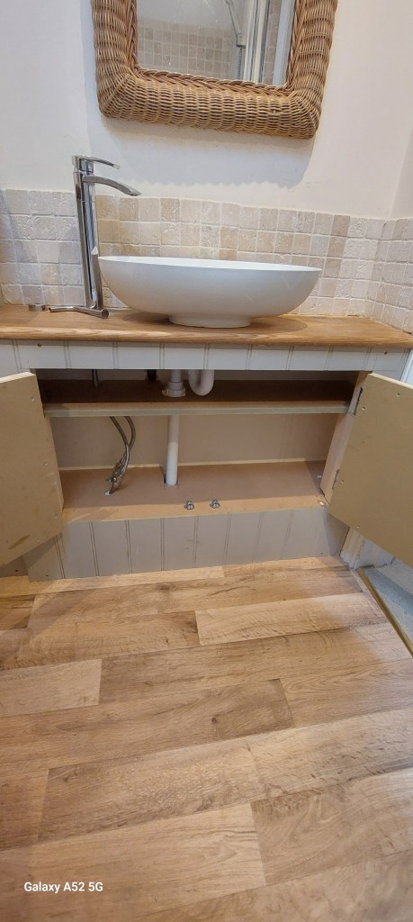 A modern bathroom sink with a white vessel bowl on a wooden countertop, complemented by a stylish faucet. Below, cabinet doors are open to reveal plumbing. The wall is adorned with mosaic tiles, and a woven mirror hangs above.