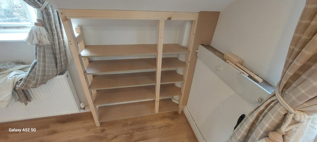 A wooden shelf unit with several shelves installed in a room, next to a window with a light-colored curtain. There is a radiator and some fabric draped nearby.