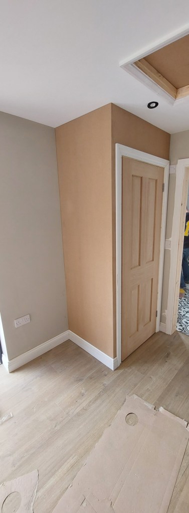 Interior corner of a room showing a light gray wall and a plywood wall with a wooden door, with flooring exposed and construction materials scattered on the floor.