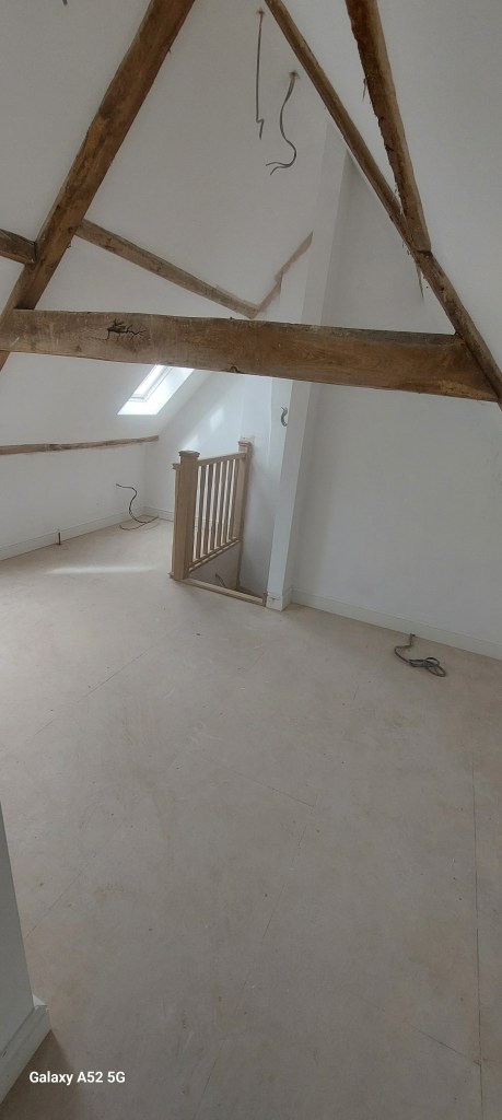 Interior view of an unfinished attic space featuring wooden beams, a staircase, and a window.