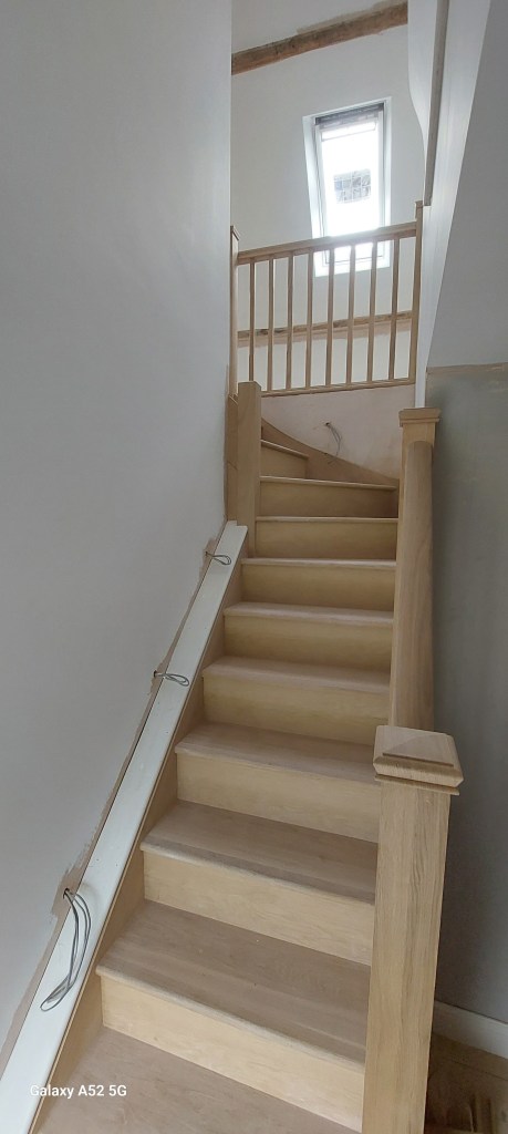 A well-lit wooden staircase leading up to a landing with a window, featuring a simple railing and smooth steps.