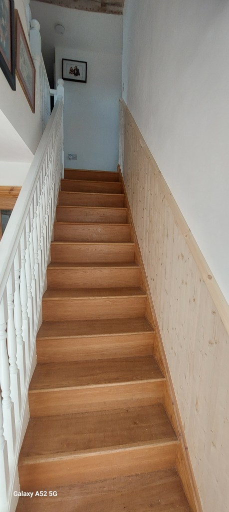 A wooden staircase leading upwards, with white railings and light-colored walls adorned with framed pictures.