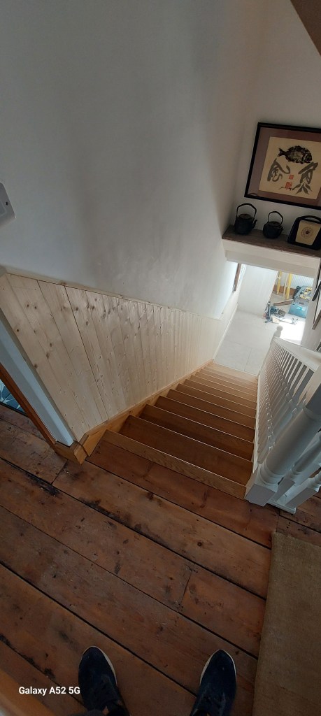 A view looking down a staircase with wooden steps, featuring wooden wall paneling and a light-colored wall. Art is displayed on a shelf at the top of the stairs.