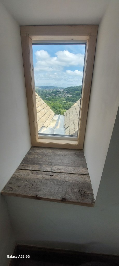 A wooden window frame with a view of green hills and blue sky, featuring fluffy clouds, from an interior space.