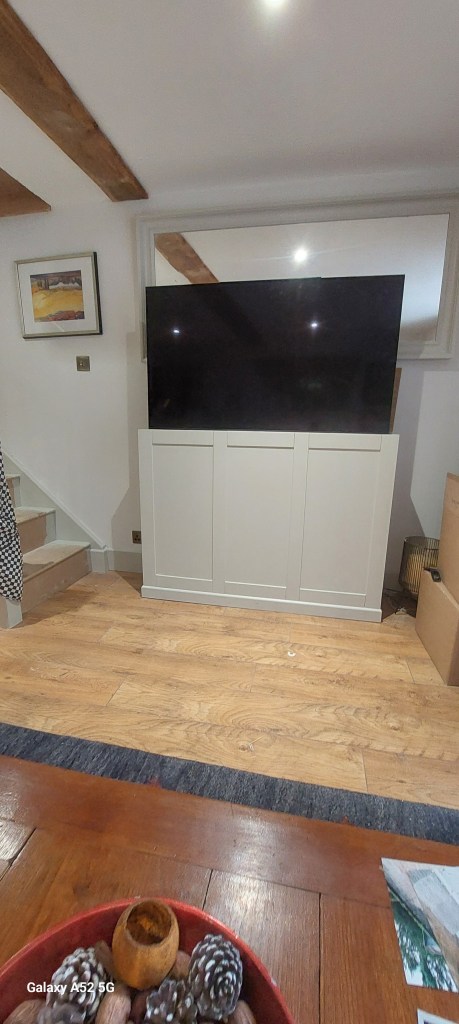 A living room with a television mounted on a white cabinet, wooden flooring, and a bowl of pinecones on a table in the foreground.