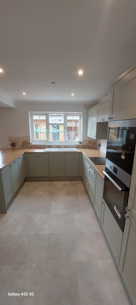 A modern kitchen with light green cabinetry, a wooden countertop, and stainless steel appliances. The space features ample lighting, a large window, and a minimalistic design.