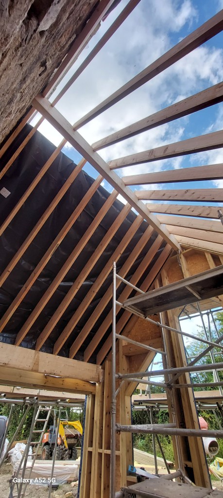 View of a construction site showing wooden beams and scaffolding, with a blue sky visible through the open roof structure.