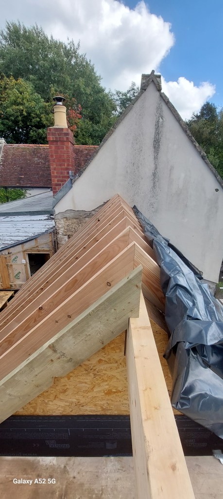 View of a roof construction with wooden beams, showcasing a chimney and a partially completed structure under a clear sky.