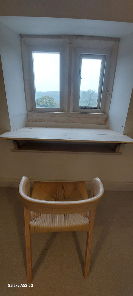A view of a minimalist workspace featuring a simple wooden chair at a small desk positioned beneath two windows, with a scenic landscape visible outside.
