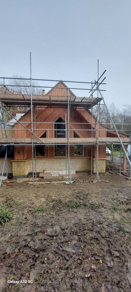 A wooden house under construction, featuring scaffolding around it. The structure has a triangular roof and large windows, with a muddy ground surrounding the building site.
