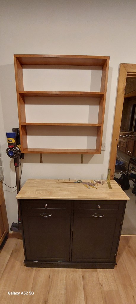 A wooden shelving unit mounted above a dark cabinet with a light-colored tabletop. The cabinet has two doors and is positioned on a wooden floor.