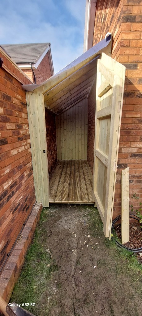 A newly constructed wooden shed with an open door, showing the interior and wooden flooring, situated between brick walls in a residential area.