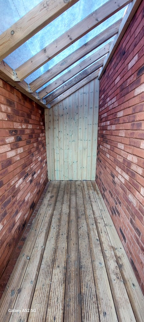 Interior view of a wooden structure with brick walls and a sloped roof, showcasing wooden flooring and a wooden back wall.