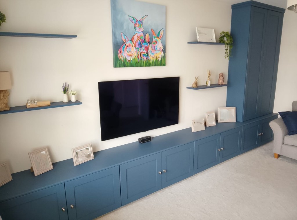 A living room with a blue media unit below a flat-screen TV, decorated with framed pictures and small decorative items on shelves. Above the TV, there is a colorful painting of rabbits.