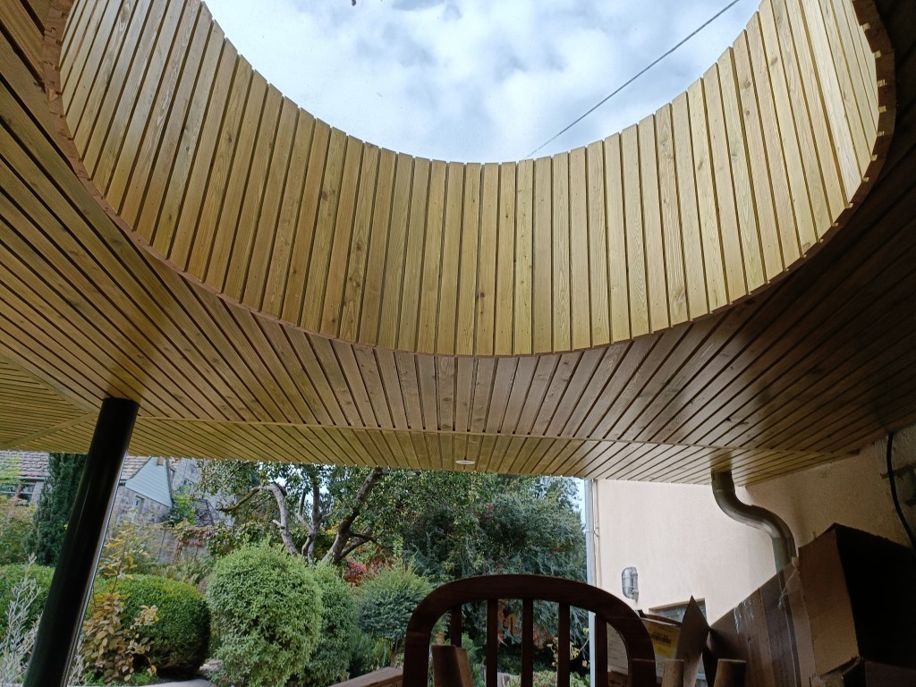 View of a circular wooden ceiling with an opening for natural light, surrounded by greenery in the background.