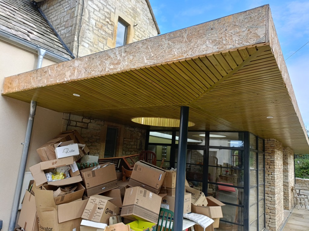 Exterior view of a house featuring a modern roof with wooden paneling, stacked with cardboard boxes and situated next to stone walls.