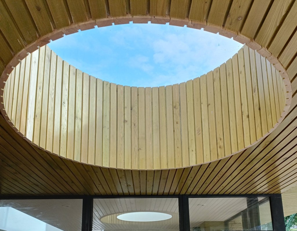Interior view of a wooden structure with a round skylight revealing blue sky, surrounded by wooden paneling.