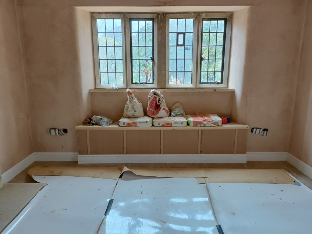 Interior view of a room under renovation, featuring incomplete walls in a soft pink hue, a row of windows with decorative panes, and a wood bench with construction materials on top.