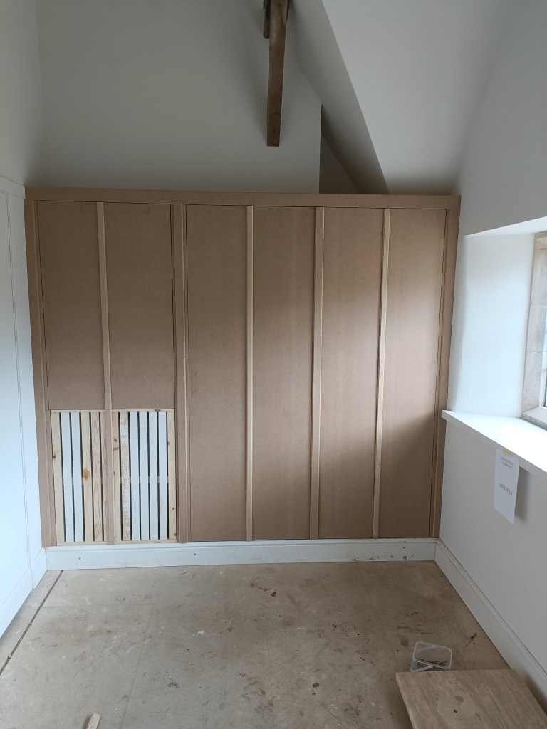 Photo of an unfinished interior wall featuring MDF panels with vertical lines and a small wooden vent cover at the base. The room has sloped ceilings and a window providing natural light.