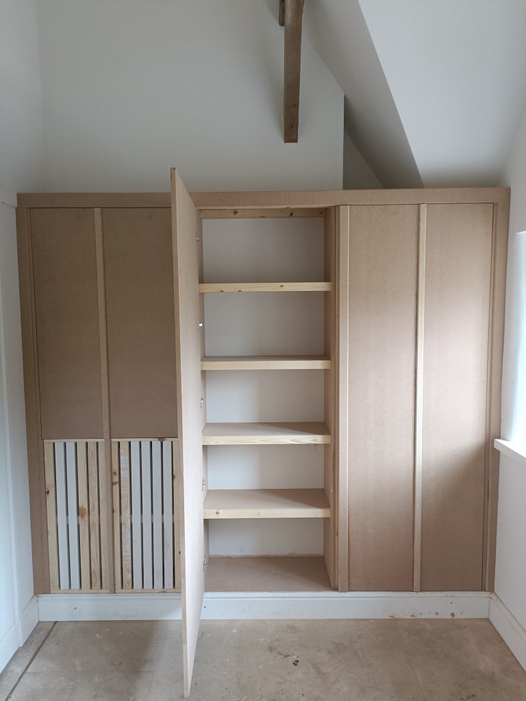 Interior view of a wooden cupboard with an open section revealing shelves. The cupboard features a simple design, with a combination of panels and slatted wood, set against a wall with a sloped ceiling.
