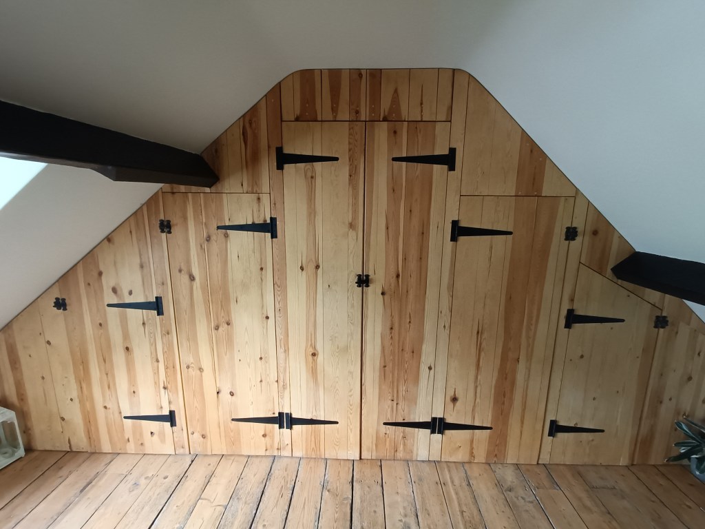 A wooden wall in an attic featuring multiple doors and decorative black hinges, with a slanted upper edge and wooden flooring.