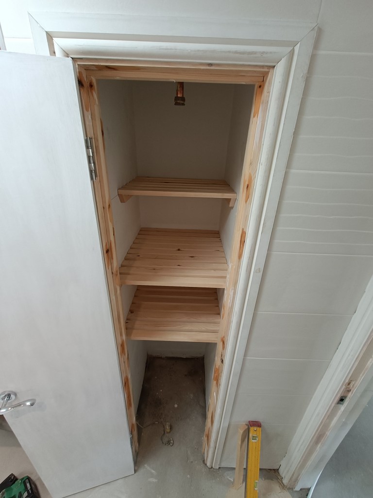 A view of an unfinished small closet, featuring wooden shelves and a door, with construction materials visible on the floor.