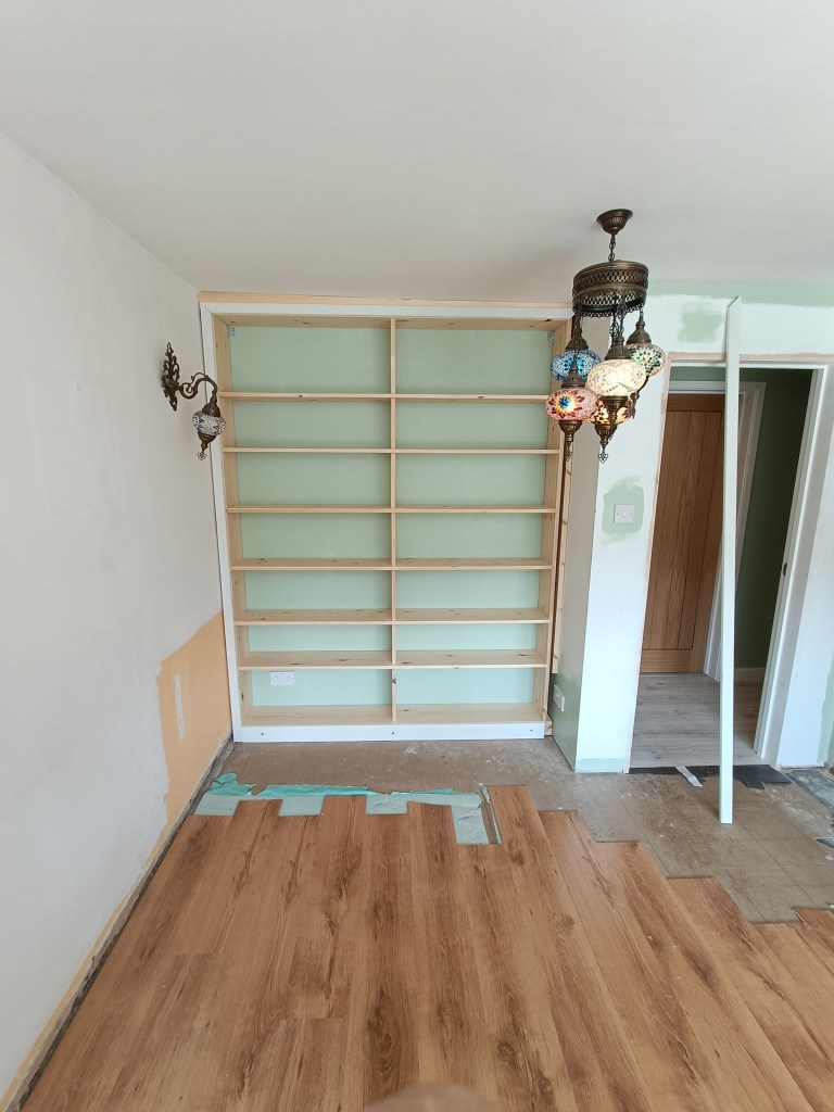 A partially finished room featuring a wooden floor, an unfinished wall with a wooden shelf structure, and colorful hanging lights.