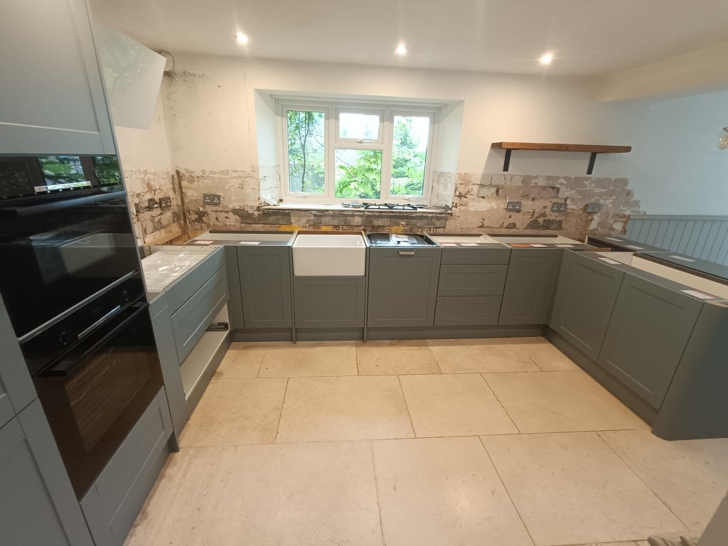 A modern kitchen with grey cabinets, featuring a farmhouse sink and an oven. The space is undergoing renovations, with exposed walls and natural light coming through a window.
