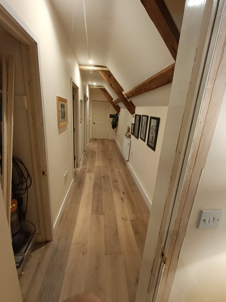 A narrow hallway with wooden flooring, featuring exposed beams on the ceiling and several doors along the walls, some with artwork displayed.