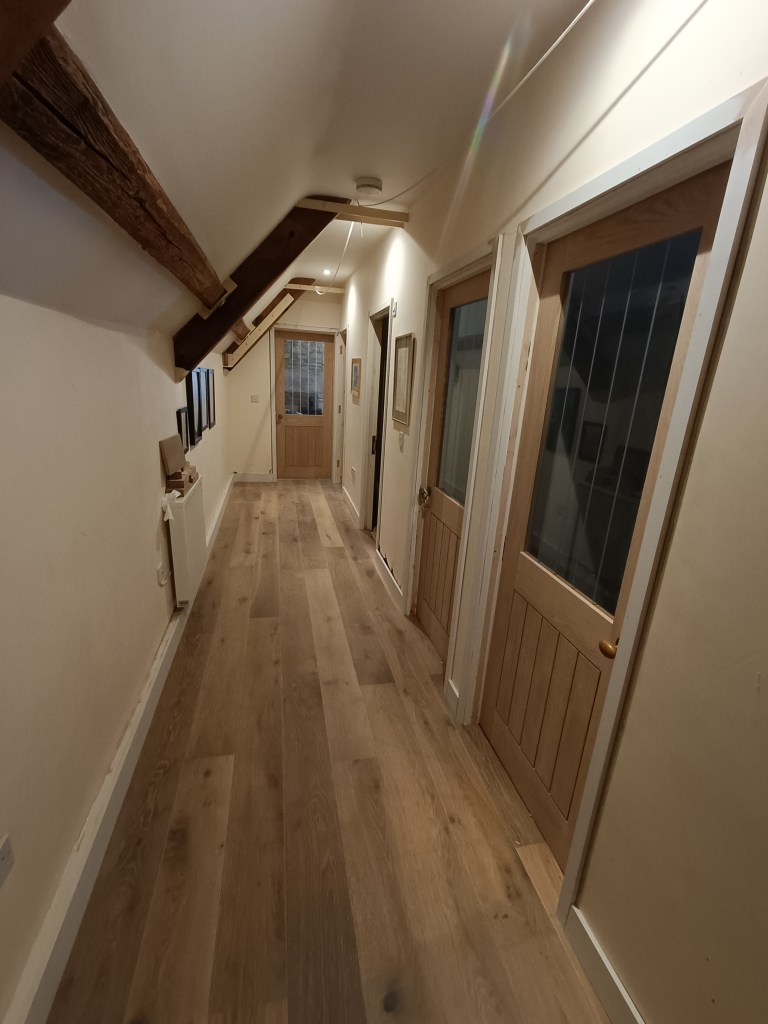 A well-lit hallway featuring wooden flooring and wooden doors on both sides, with rustic wooden beams on the ceiling.