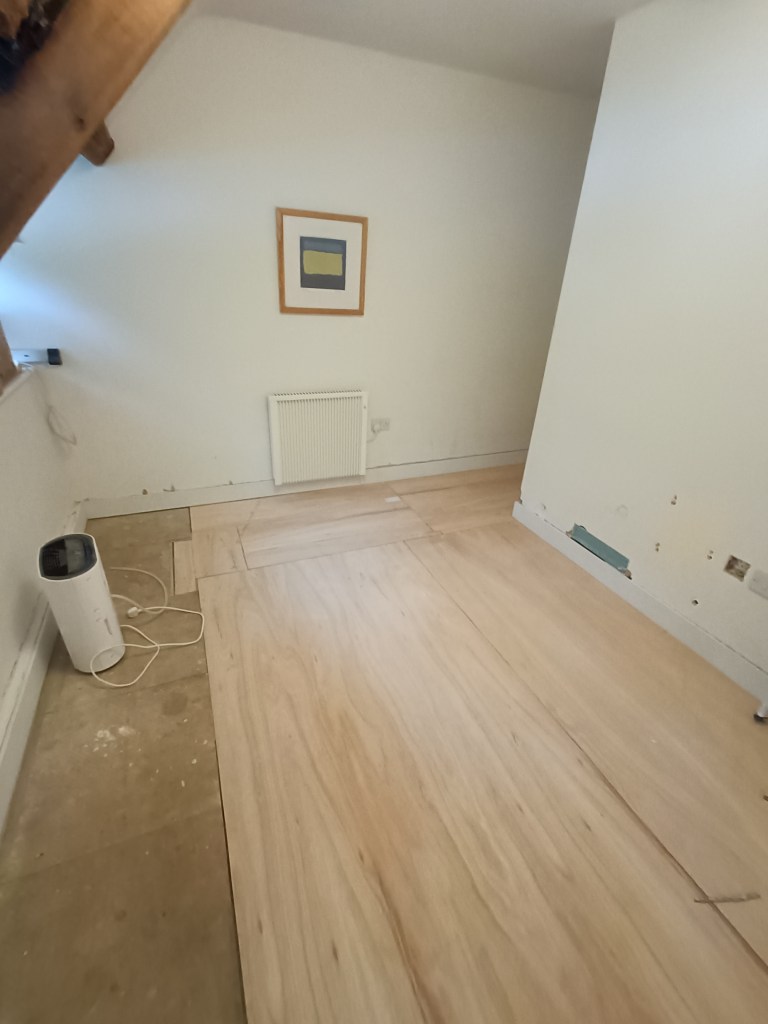 A room with wooden flooring partially installed, featuring a white wall with a framed painting. A white radiator and a small appliance can be seen on the floor.