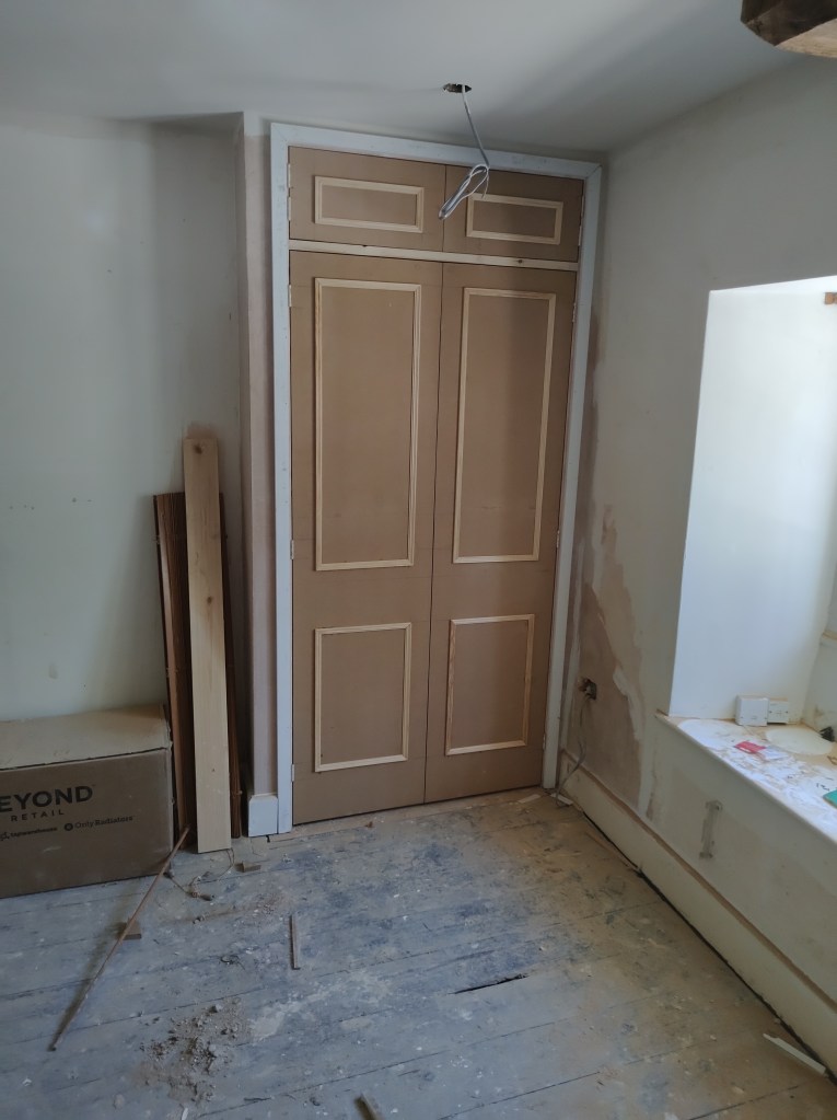 A view of a room showing a newly installed door with a plain design. The door is not yet painted and is surrounded by unfinished walls and a dusty floor with construction debris.