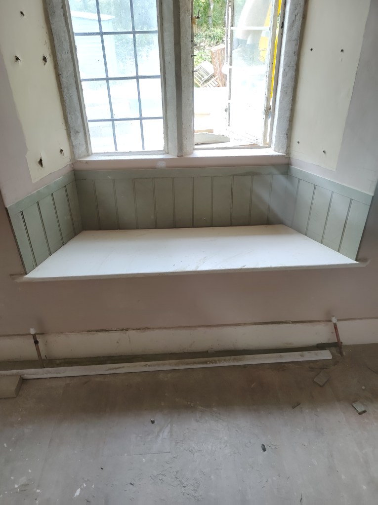 A window seat with a light green paneling below, situated in front of a large window with multiple panes, displaying an outdoor view.