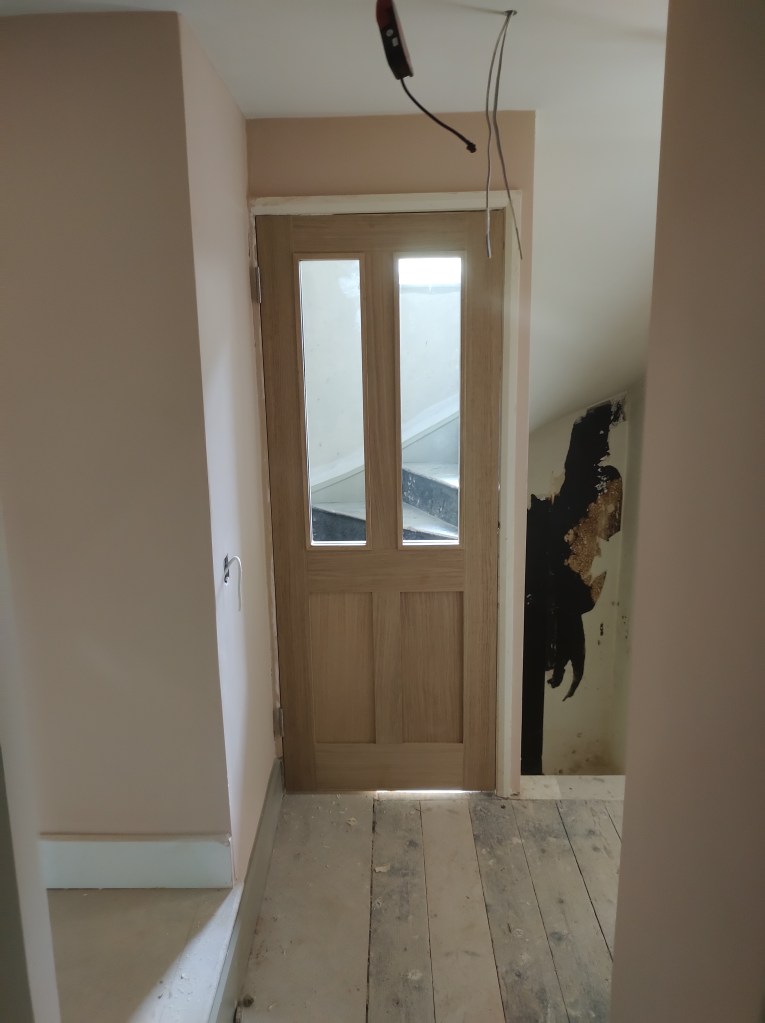 Interior view of a hallway with a wooden door featuring glass panels, light-colored walls, and exposed floorboards.