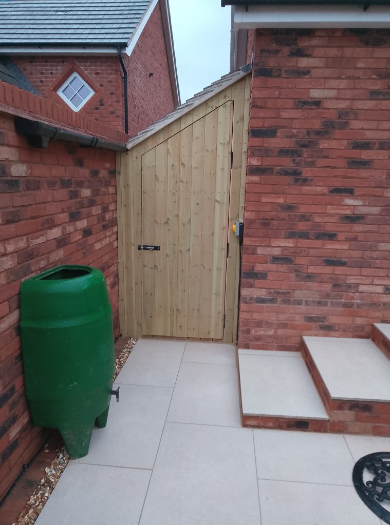 A wooden gate next to a brick wall, with a green water barrel nearby and tiles leading to a set of steps.