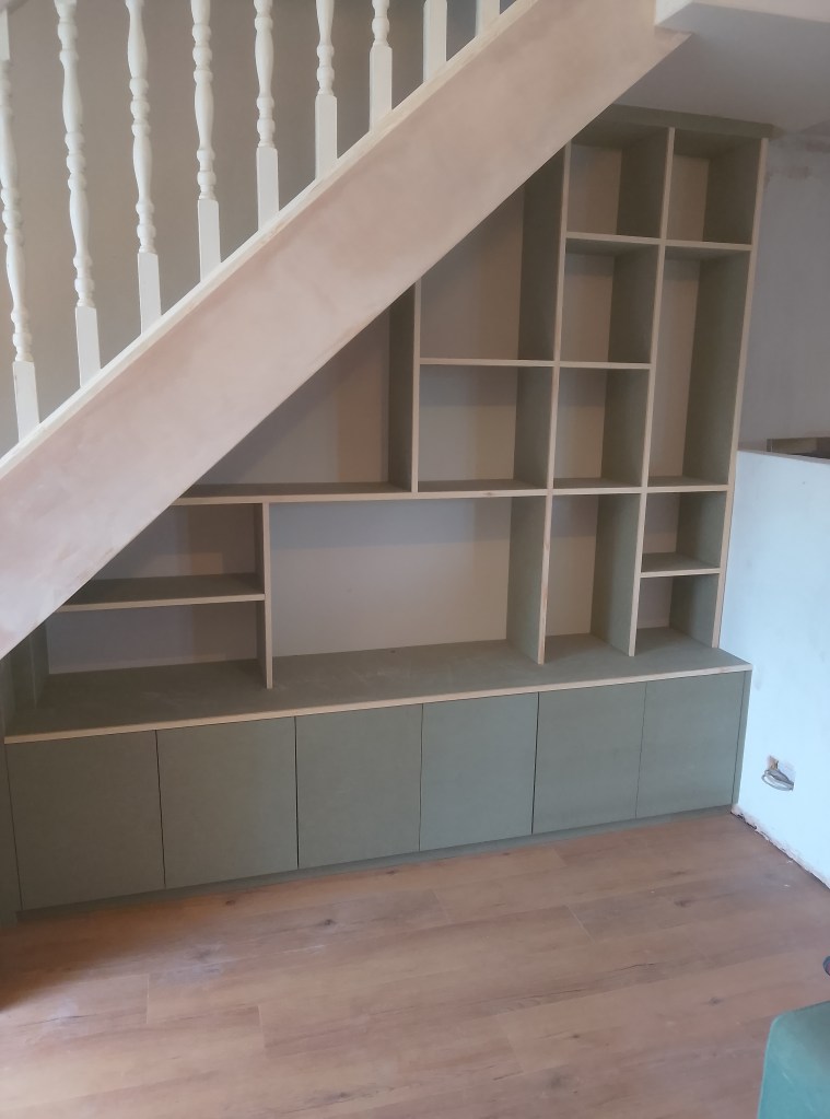 A modern wooden shelving unit installed under a staircase, featuring open shelves and closed cabinets, with a simple design and neutral colors.