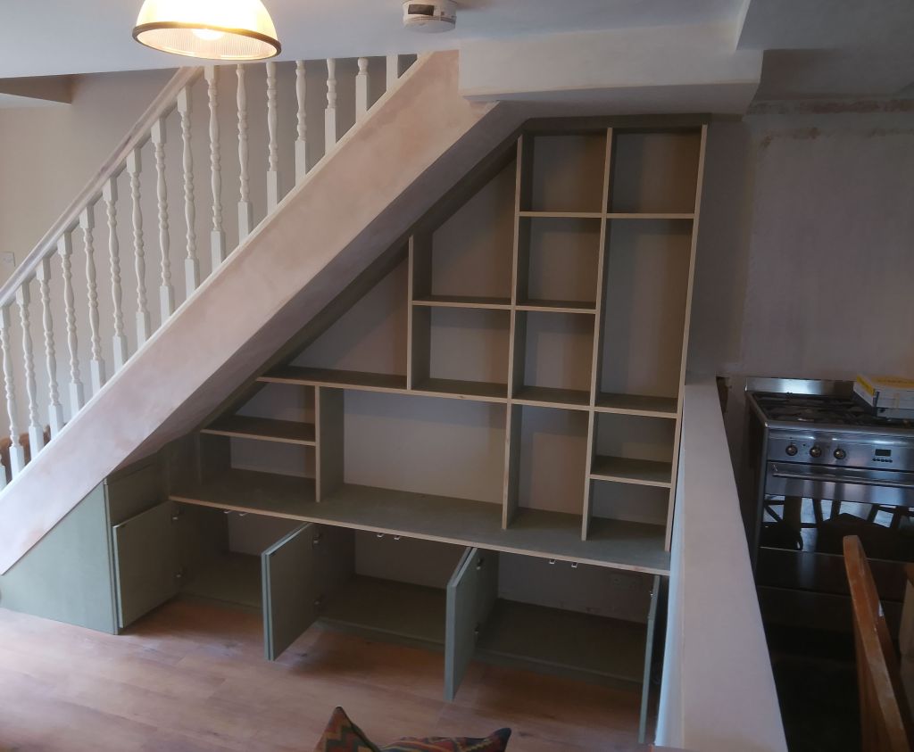A modern wooden bookshelf and storage unit built under a staircase, featuring open shelves and closed cabinets, with a staircase railing visible on the left.