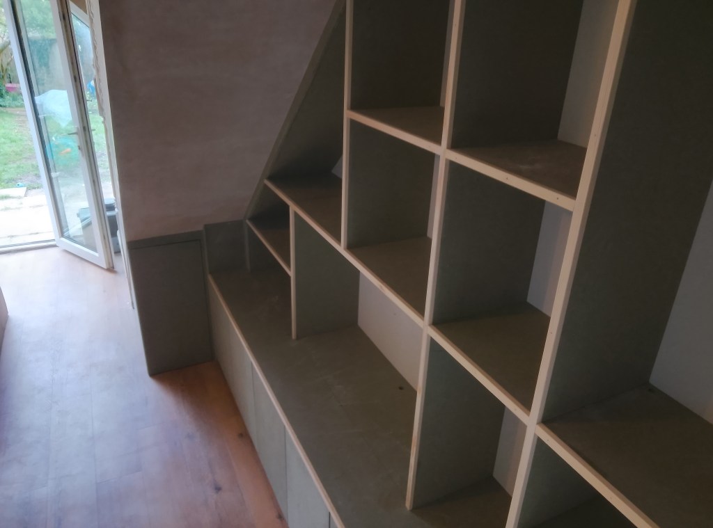 A partially constructed shelving unit in a room, with light-colored shelves and a concrete wall. A door leading outside is visible in the background, with grass and some objects in the garden.