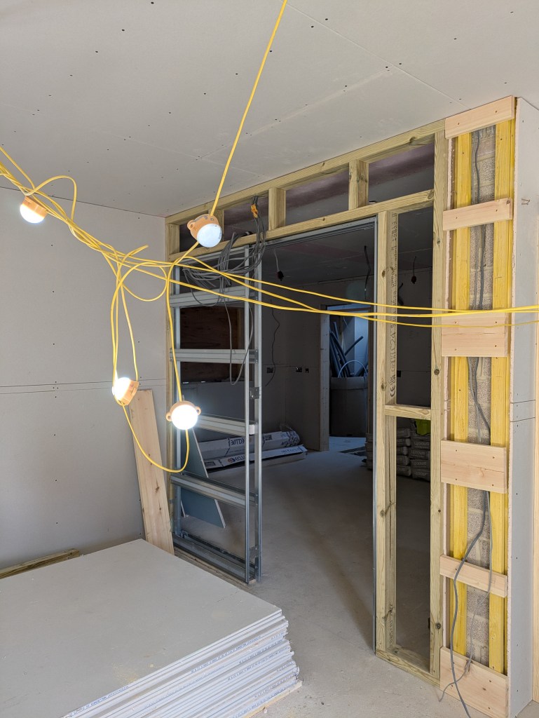 A construction site interior showing exposed drywall, bright yellow power lines, and lighting fixtures. Stacks of drywall sheets are visible in the foreground with an unfinished doorway framed in wood on the right.