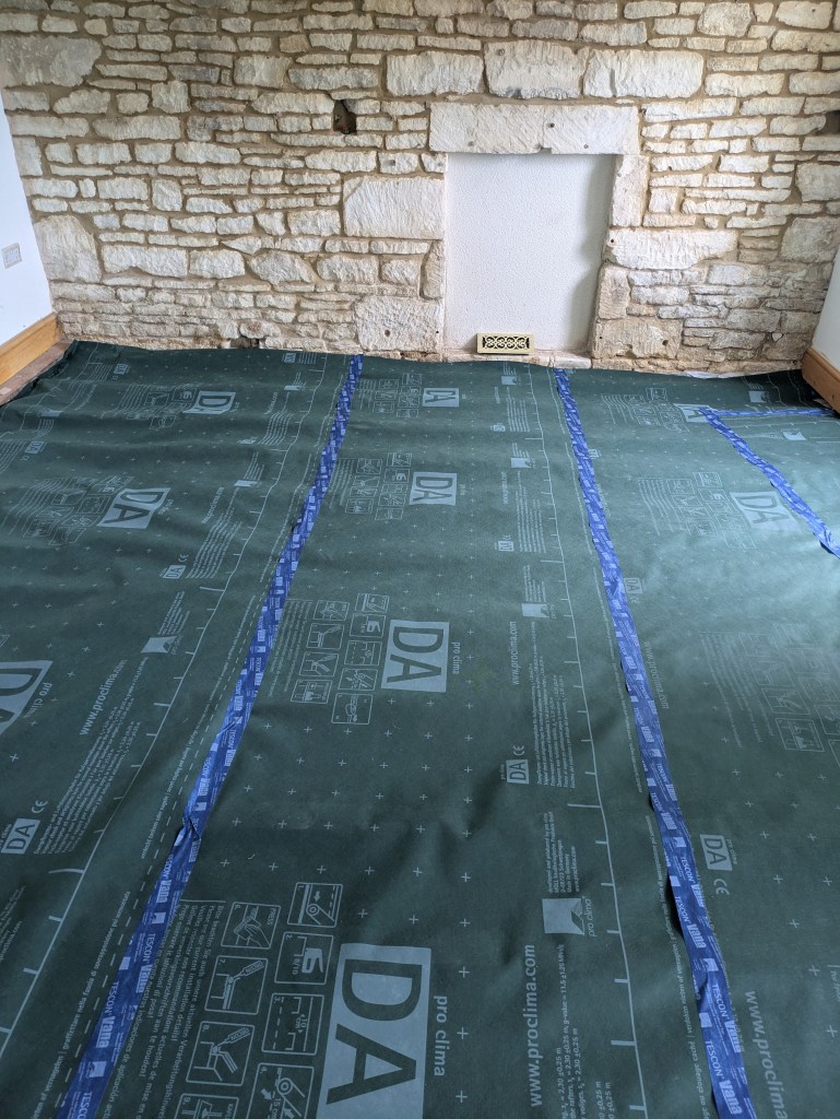 A floor covered with a green vapor barrier roll featuring markings, placed in front of a stone wall and a fireplace opening.